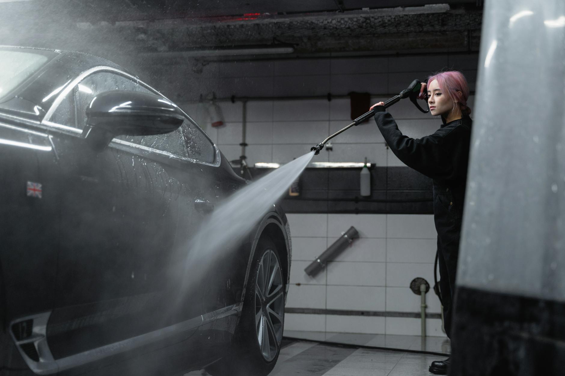 Woman power washing a black car in an indoor garage using a high-pressure washer.